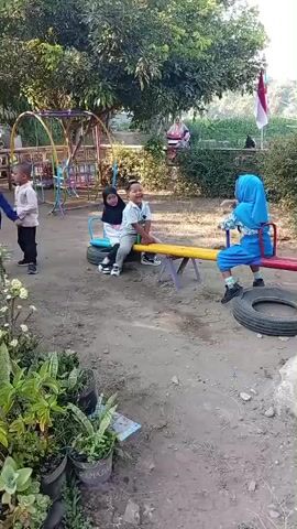 The video opens on a children's playground with a seesaw as the central focus. Four young children are present. One child, wearing a blue hijab and dress, is seated on the right side of the seesaw, which is supported by tires. Another child, wearing a white shirt and gray pants, and a child in a black hijab and gray pants, are seated on the left side of the seesaw. A fourth child, wearing a light-colored long-sleeved shirt and black pants, stands to the left of the seesaw. Behind them, a swing set is visible, with another child and an adult near it. The playground appears to be an outdoor area with dirt ground and trees in the background. The sun is shining, casting shadows. The video pans slightly, revealing more of the playground and surrounding vegetation. The overall atmosphere is one of a typical day at a playground.