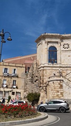 The video shows a tall stone building in the city of Cagliari, Italy. The building is decorated with windows, crosses, arches, and other architectural details. People are seen walking around on the steps leading up to the building. A banner hangs from the top of the building, advertising an event that runs from February 28 to July 6, 2024. A white car is parked on the street next to the building.