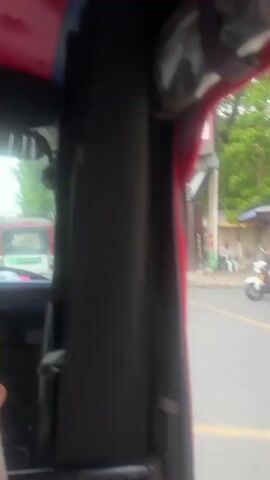 The video provides a first-person perspective from inside a vehicle, likely a tuk-tuk or similar open-air transport. The view is primarily focused on the interior, with a black fabric panel dominating the right side of the frame. To the left, a glimpse of the outside world is visible through a window. A blue lanyard with a white tag displaying a QR code and text "ARA PAY" hangs prominently in the foreground. The tag is attached to a metal bar. Beyond the bar, a person's arm and shoulder, wearing a light-colored shirt, are visible, suggesting the cameraman is a passenger. Through the window, a street scene unfolds with trees lining the road and other vehicles, including a white and red bus and several motorcycles, in motion. The lighting suggests it is daytime, possibly overcast, with sunlight filtering through. The overall impression is that of being transported through an urban or semi-urban environment.