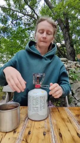 A woman with blonde hair in braids, wearing a teal hoodie and patterned pants, is sitting at a wooden table outdoors, preparing to light a portable camping stove. She is holding the stove with both hands, adjusting it on top of a white gas canister labeled with black text. To her left, a metal mug sits on the table. The background shows a wooden bench, a stone wall, and lush green trees. The woman appears to be focused on the stove.