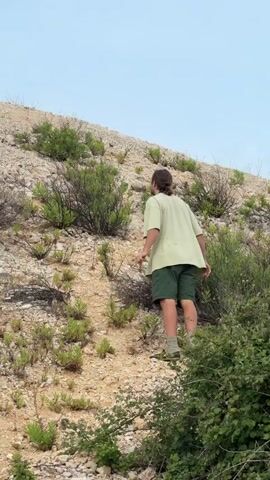 A person is climbing up a small, sparsely vegetated hill towards a large wind turbine. The person is wearing a light green t-shirt and dark green shorts. They pause with their arms outstretched at one point. The wind turbine has the letters "SPIC" and a logo on it. The sky is pale blue with some clouds.