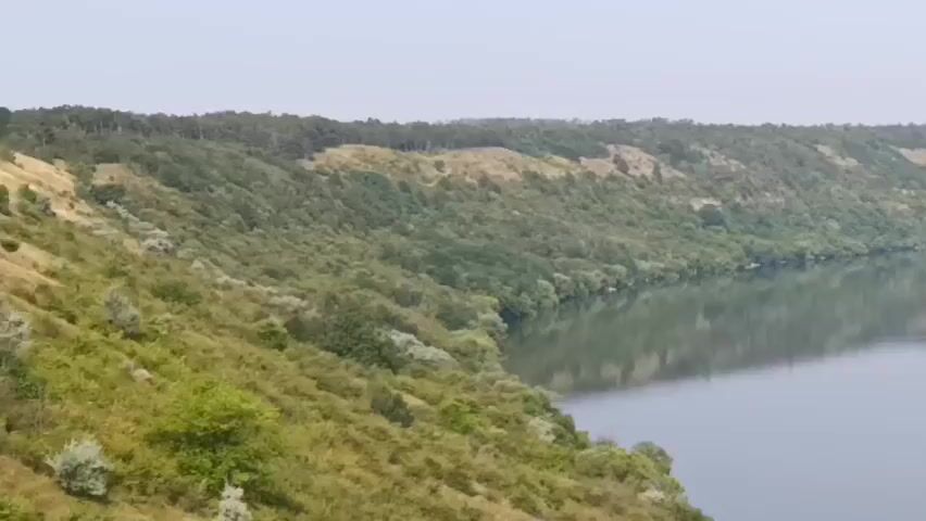 A wide shot shows a tranquil body of water reflecting the sky. On the left side, a lush, green, and hilly landscape slopes down to the water's edge, covered with trees and shrubs. The terrain continues along the curve of the water, with patches of lighter earth visible among the greenery. The sky is a pale, hazy blue.