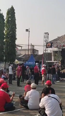 A group of people are gathered in an outdoor area, possibly a street or plaza. The foreground shows several individuals sitting on the ground, facing away from the camera. They are wearing red shirts and some have red hats. In the midground, a group of people are standing. Some are wearing red shirts and white hats, and some are wearing red and white striped shirts. One person in the midground is holding a red object with a cartoon face on it. Further in the background, there is a stage structure with a banner, a blue tent, and buildings. Trees are visible on the left side of the frame. The sky is clear and bright, indicating daytime. One person in the midground is walking backward.