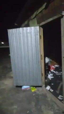 The video begins by showing the inside of a dilapidated structure. A large metal roll-up door is partially open on the left, revealing a dark interior filled with debris and trash. To the right, a doorway opens into the same dark, trash-filled space. The camera then pans to the right, showing the exterior of a large, rusted metal dumpster with blue paint. The scene transitions to an outdoor area at night. The ground is covered in dirt, gravel, and some sparse vegetation. In the distance, there are faint lights, suggesting the presence of buildings or streetlights. The camera moves further into the dark outdoor environment, showing more of the uneven ground and some piles of what appear to be construction materials, possibly lumber or bags. The camera then turns and shows the side of a brick wall, possibly part of a building. The final frames show more of the dark outdoor area with distant lights. The video appears to be a shaky, handheld recording.