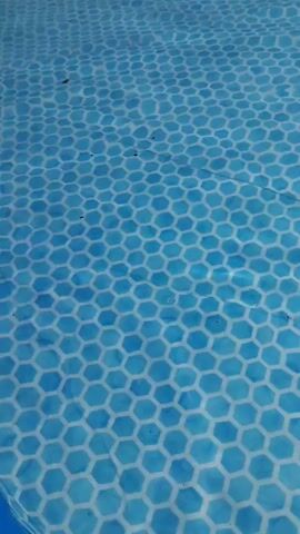 A hand with a tattoo of a vine on the wrist and palm is shown dipping into blue water. The water has a hexagonal pattern on the bottom. The hand moves through the water creating ripples.