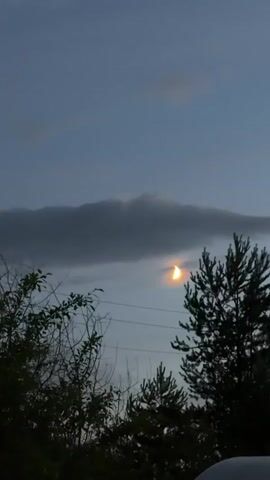 A video shows a cloudy sky during twilight with a crescent moon visible through a break in the clouds. The moon is partially obscured by a dark cloud, and a halo effect with rainbow-like colors surrounds it. Trees are visible in the foreground, silhouetted against the sky. Power lines are also present in the frame. The camera is stationary.
