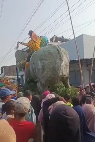 A large, elephant-shaped float covered in light-colored fabric is central to the scene. A person dressed in yellow and black is seated on top of the float, holding onto a pole. The float is surrounded by a crowd of people of various ages, some wearing hats and masks. The environment appears to be an outdoor street or parade route, with buildings and utility poles visible in the background. The sky is bright, suggesting it is daytime. There are green plants decorating the base of the float. The crowd is observing the float and its rider.