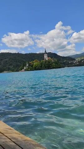 The video opens with a shot of the crystal-clear, turquoise waters of Lake Bled in Slovenia. The camera is positioned low, close to the water's surface, revealing the sandy bottom beneath. Ripples gently disturb the surface, reflecting the bright blue sky dotted with fluffy white clouds. In the distance, across the lake, the iconic Bled Island is visible, with its picturesque church featuring a tall, slender spire. The island is nestled amidst lush greenery and surrounded by the calm waters of the lake. Further in the background, rolling hills and mountains form a majestic backdrop, with a few buildings scattered along the shoreline. The scene is bathed in the warm light of a sunny day, suggesting a pleasant time for outdoor activities. A traditional wooden boat, known as a pletna, is seen gliding across the water, adding to the idyllic charm of the location. The overall atmosphere is one of tranquility and natural beauty, evoking a sense of peace and wonder. There is no visible text in this media file.