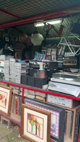 The video shows an outdoor flea market stall filled with various items. Stacks of CDs and vinyl records sit on the upper shelves alongside vintage audio equipment like turntables and amplifiers. Several framed art pieces rest on the lower shelves. Above, kitchenware like pots and pans hang from the ceiling. The stall has a red-framed structure and a green wire mesh roof. The background outside the stall is blurred, suggesting an outdoor setting.