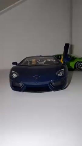 The video shows two toy cars on a white shelf against a white wall. The toy car in the foreground is a dark blue Lamborghini with its driver side door open. Behind the blue car is a green Lamborghini.