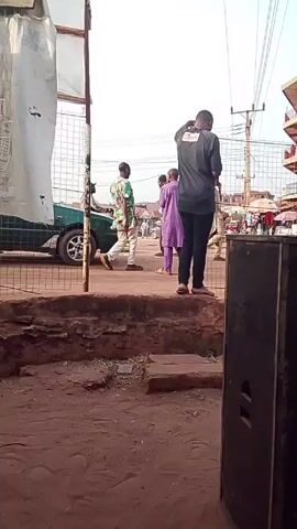 The video shows a street scene with people walking and a parked car. A person in a gray shirt stands on a raised platform, possibly taking pictures or recording a video. Other people walk past, some carrying children. There is a building in the background and power lines overhead. A speaker is visible in the foreground. The ground is reddish-brown dirt.