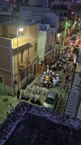 A procession of people walks down a narrow street lined with buildings. In the foreground, a dark, patterned surface is visible. The procession includes individuals in white robes, some carrying a large, ornate casket adorned with flowers, and others in a band uniform playing instruments. Several cars are parked or moving slowly in the street. The scene appears to be at dusk or in the evening, with artificial lights illuminating the area.