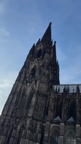 This video shows a low-angle, panning shot of a large, gothic cathedral. The cathedral is made of dark stone and features intricate architectural details, including spires, arched windows, and buttresses. The sky above is a clear blue with a few wispy clouds. In the lower part of the video, a crowd of people is visible walking in front of the cathedral. Some of the people are blurred, suggesting movement. The lighting suggests it is daytime, possibly late afternoon. There is no audio present in the video.