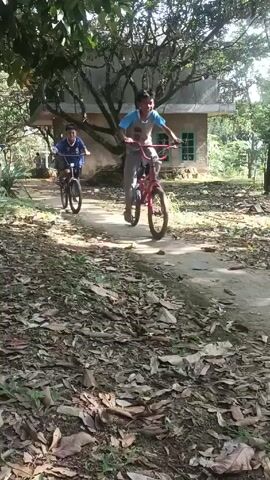 Two young people ride bicycles on a dirt path covered in dry leaves, past an old house with light green window frames.  Trees stand around the house and on either side of the path. One person is riding a red bicycle. The other is on a dark-colored bicycle. The camera is stationary and low to the ground.