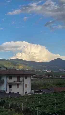 The video captures a scenic view from a moving vehicle, likely a train, in Nave San Rocco, Italy. The dominant subject is a dramatic, towering cumulonimbus cloud formation illuminated by the setting sun, creating a striking contrast against the clear blue sky dotted with smaller clouds.

The scene unfolds with a landscape of rolling green hills and vineyards, interspersed with small clusters of houses and buildings, suggesting a rural or semi-rural setting. In the foreground, lush green foliage, likely vineyards, blurs due to the motion, framing the more distant view. Further back, several houses with red-tiled roofs are visible, along with what appears to be a sports field or park area with green turf and floodlights. The background is dominated by the imposing mountain range, where the massive cloud has settled, catching the golden light of the late afternoon or early evening.

The overall mood is one of tranquility and natural beauty, amplified by the grandeur of the sky and the picturesque Italian landscape. The time of day is clearly indicated by the warm, directional light bathing the clouds and the mountains, suggesting either sunrise or sunset. The weather appears dynamic, with the large cloud potentially indicating an approaching or dissipating storm.

There are no visible people or specific objects that stand out as central to the action, beyond the natural elements and the infrastructure like houses and sports facilities. The motion blur of the foliage in the foreground and the gradual shift in the landscape as the vehicle moves are key indicators of the activity—travel.

A faint blue and white sign is partially visible on the right side of the frame at the 00:00 mark, but no discernible text can be read. The focus remains on the natural spectacle and the serene Italian countryside.