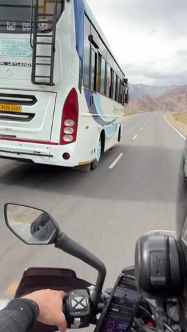 The video begins with a first-person perspective of a person riding a motorcycle on a paved road. A large white bus with blue and white accents drives alongside the motorcycle on the left. The road is surrounded by barren, mountainous terrain with a cloudy sky overhead. The motorcycle rider's hand is visible on the handlebars. As the video progresses, the motorcycle pulls ahead of the bus, and the road continues through the mountainous landscape. The bus is then seen in the distance behind the motorcycle, continuing down the road. The motorcycle appears to be equipped with a phone mount displaying navigation or other information.