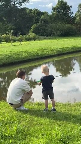 The video shows a man and a child fishing by a pond. The man is crouched down, wearing a white t-shirt and beige shorts. The child is standing, holding a fishing rod, and wearing a dark t-shirt and shorts. The pond is surrounded by lush green grass and trees. In the background, there are more trees and some bushes. The water in the pond is calm and reflects the trees and sky. Later in the video, a calico cat appears in the grass to the left of the pond.