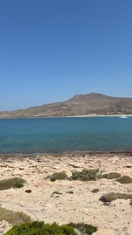 The video shows a scenic view of a rocky beach with sparse vegetation in the foreground. The sea is a vibrant turquoise, with a white sailboat anchored in the distance. Behind the sea, a large, barren mountain dominates the horizon. The sky is a clear, cloudless blue. The camera appears to be stationary, capturing a still image of the landscape. The timestamp indicates that the video is approximately 4 seconds long.