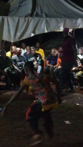 The video shows a person in colorful clothing dancing on grass in front of a seated audience under a white tent. The dancer, wearing a red, black, and yellow outfit, moves energetically while the audience, mostly men, watches and some gesture. The tent is lit from within, and the setting appears to be outdoors at night. A person in a maroon shirt stands to the right, partially obscuring the view.