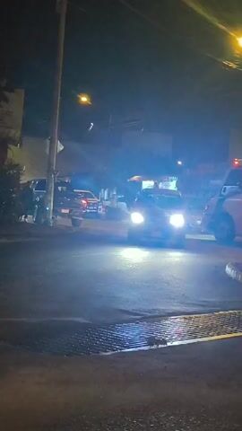 The video depicts a night scene on a street with several vehicles, streetlights, and a food stand. A dark-colored vehicle approaches the intersection with its headlights shining brightly. The food stand, illuminated with festive lights, has several people gathered around it. A white van with flashing lights is parked on the side of the road.  The street surface is wet and a storm drain can be seen in the foreground. A power pole is also visible.