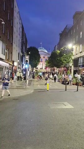 A street scene at dusk shows a building with a domed roof at the end of a street lined with buildings. There are people walking and sitting on the street, and a cyclist is riding a bicycle. Traffic lights are visible, and streetlights are illuminated. The sky is a deep blue, indicating that it is evening.