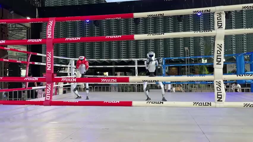 The video shows two robots in a boxing ring. The robot on the left is wearing red boxing gloves, while the robot on the right is wearing white gloves. Both robots are white and appear to be humanoid. They are standing opposite each other in the ring, with ropes dividing the space. The background shows a structure with many vertical elements, possibly a building or a stage set. There are some people visible in the background, as well as lighting fixtures. The overall environment appears to be outdoors or in a large open space, possibly an event or a demonstration. The robots are posed as if ready to engage in a boxing match.