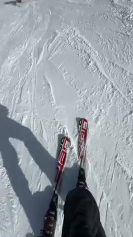 The video shows a first-person perspective of someone skiing down a snow-covered slope on a sunny day. The skier's skis are visible at the bottom of the frame, and their shadow is projected onto the snow.  Other skiers can be seen further down the slope. A ski lift runs along the slope. The background shows a view of trees and distant land under a blue sky with scattered clouds.