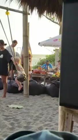 The video shows a group of people eating at a table on a beach. In the center of the table is a large roasted pig. A man wearing a blue shirt and a hat is decorating the pig with a yellow, cone-shaped object.  The table is also decorated with red peppers and green leaves. Behind the table is the ocean, and the sun is setting in the background. There are black beanbag chairs around the table. To the left is a wooden structure. A child walks to the left of the scene.