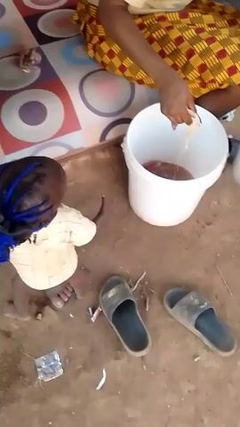 A person is sitting on the floor in a yellow and red checkered skirt, transferring pieces of what appears to be raw chicken from a white bucket into a metal pot. A small child stands nearby. The floor is a dirt surface. Two sandals lie on the floor. There is a patterned mat and other miscellaneous items in the background.