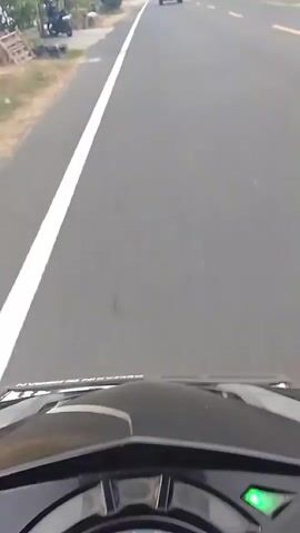 The video shows a first-person perspective from a motorcycle or scooter traveling on a paved road. The road has a white line on the left and a yellow line in the center, indicating two lanes. In the distance, several white cars are visible, some ahead and some approaching. The environment appears to be rural or suburban, with vegetation and utility poles along the side of the road. The sky is overcast. The motorcycle's dashboard, showing a green indicator light, is partially visible at the bottom of the frame. The camera is moving forward at a steady pace.