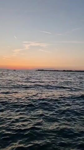 This is a video taken from a boat at sea. The time of day appears to be sunset or sunrise, as indicated by the warm orange and pink hues in the sky. The water is dark and choppy, with small waves. In the distance, a silhouetted coastline is visible with a cluster of buildings, possibly a town or city. A small boat or ship can be seen on the far left of the horizon. The sky is a gradient of pale blue at the top, transitioning to soft orange and pink near the horizon. There are a few wispy white clouds, one of which appears to be a contrail from an airplane. The overall mood of the video is peaceful and serene, evoking a sense of calm and natural beauty. The location is identified as Zadar, Croatia. There is no visible text in the video.