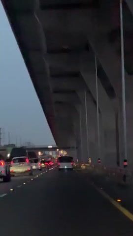 The video shows a view from a vehicle driving on a highway at dusk. The highway is elevated, with a large concrete structure above, likely an overpass or bridge. The sky is a dark, overcast blue. Traffic is visible ahead, with cars in multiple lanes moving slowly, suggesting congestion. The headlights and taillights of the vehicles are illuminated. Yellow reflective markers are visible along the right side of the road, and orange and white traffic cones are also present. The overall atmosphere is one of a busy commute during evening hours.