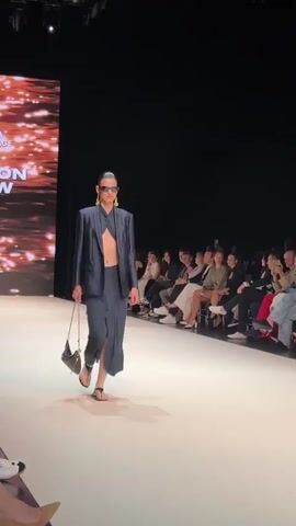 The video shows a fashion show. A female model walks down a runway wearing a navy blue suit with a cutout top. She has on large sunglasses and gold earrings. She is carrying a black handbag. A large screen with a blurry, glittering image is visible in the background. Audience members are seated on the right side of the runway, watching the model.