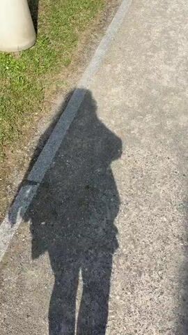 The video shows the shadow of a person walking on a paved surface. The shadow is cast by the sun from the right side of the frame, creating a long shadow that stretches across the path. The person is wearing a light-colored top and dark pants, and their hair appears to be long. They are holding a phone in their right hand, which is likely being used to record the video. To the left of the path, there is a grassy area with some plants. The path is bordered by a strip of lighter-colored material, possibly granite. In the background, a metal grate is visible on the edge of the path. The video is shot from a downward angle, looking at the shadow on the ground. The lighting suggests it is daytime, likely sunny.