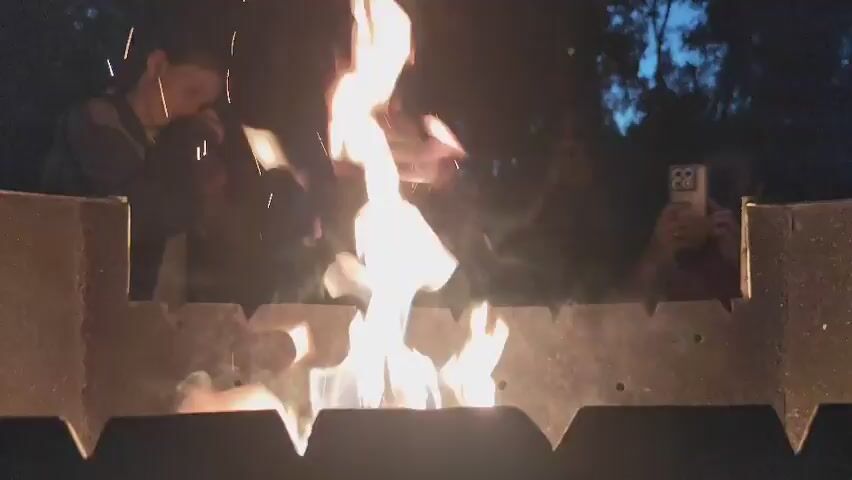 The video captures a nighttime scene centered around a roaring bonfire within a metal grill. The flames are bright and dynamic, casting a warm glow on the immediate surroundings. Several people are visible in the background, silhouetted against the fire and the darkening sky. Their faces are not clearly discernible due to the low light and distance, but their presence suggests a gathering or social event.

One individual on the far right is holding up a smartphone, appearing to record or photograph the fire. The surrounding environment suggests an outdoor setting, possibly a backyard or a park, with trees visible in the dim light behind the people. The time of day is clearly nighttime, indicated by the darkness of the sky. There are no explicit details about the weather, but the presence of a bonfire suggests it's likely cool enough for a fire.

The overall mood is relaxed and communal, with people gathered around a source of warmth and light. The focus of the video is on the captivating flames and the shared experience of observing them. There is no visible text in the media file. The context provided indicates the location is Kolpakovo, Russia.