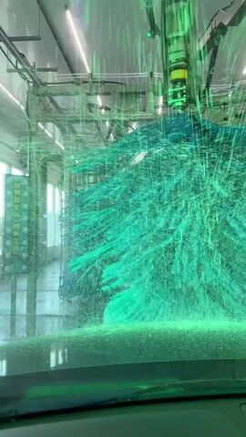 A car is moving through an automatic car wash. The camera is inside the car, looking forward through the windshield. Green soapy water and large rotating brushes are visible. The car's dashboard and steering wheel are also visible in the foreground.