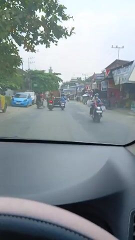 The video shows a view from inside a vehicle, looking out at a street scene. The foreground is dominated by the interior of the vehicle, specifically the dashboard and windshield. The street is paved and appears to be a two-lane road. There are several vehicles on the road, including motorcycles, a blue car, and what appears to be a small truck or utility vehicle with a trailer. Pedestrians are also visible, some on motorcycles and one walking. The buildings lining the street are varied, with some appearing to be shops or small businesses. There are trees and utility poles with overhead wires. In the distance, a building with a dome, possibly a mosque, is visible. The sky is cloudy. The overall impression is of a moderately busy street in a town or city.
