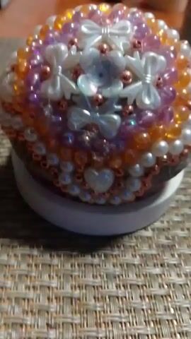A close-up shot shows a circular object covered in beads and small embellishments. The object appears to be sitting on a white base, which is placed on a textured surface that resembles a woven placemat. The embellishments include small white bows, translucent flower shapes, rose gold beads, and pearl-like beads. The colors of the beads range from light pink and purple to orange and gold. The lighting in the video is soft, and the focus is on the beaded object. There are no people or animals visible in the video. The overall impression is that of a decorative item, possibly a craft project or a small trinket box.