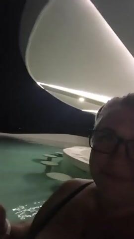 The video is a selfie-style shot of a woman with blonde hair and glasses, wearing a dark green bikini top and a necklace. She is in a pool, with a bar-like structure with circular tables visible behind her. The background appears to be outdoors at night, with some buildings and trees visible. The woman is smiling and looking at the camera. The audio features her speaking in Russian, with some background noise of water.