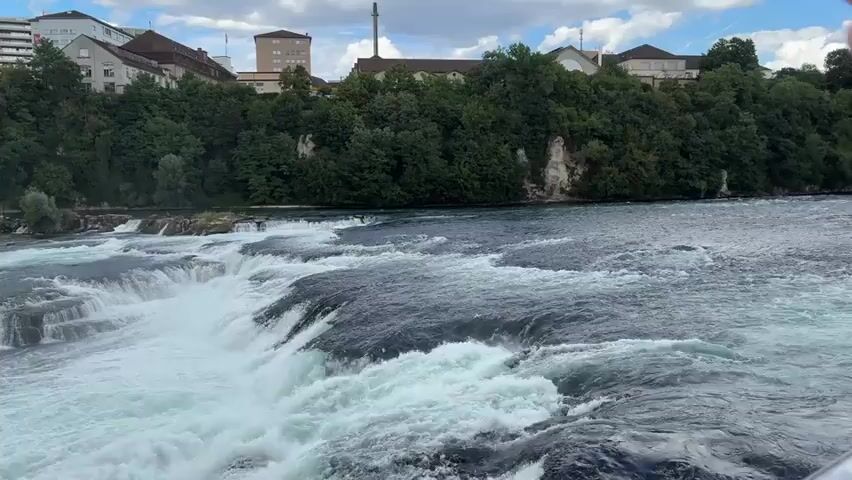 A wide shot shows a powerful waterfall with a lot of white water rushing over rocks. In the background, there are green trees and some buildings, including tall apartment buildings and smaller houses. A few people can be seen on a walkway near the shore on the left side of the frame. The sky is cloudy.