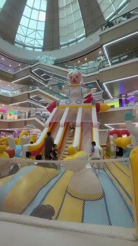 This video shows a large inflatable playground structure shaped like a rocket with an astronaut on top. Children are climbing up a ladder on the side of the rocket. In the background, a mall interior is visible, with multiple levels and shoppers.