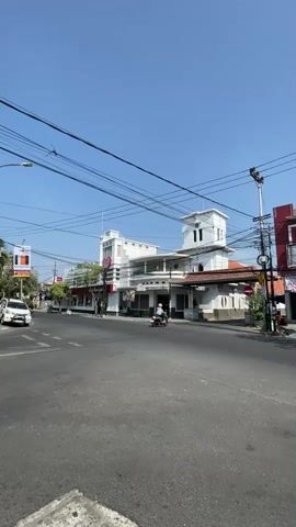 A vertical video shows a clear blue sky over a street with a building on the right side. The building is white with black trim and appears to be a historic structure with a tower. There are power lines crisscrossing the sky. A white car is parked on the left side of the street. The street itself is paved and appears to be an intersection. A motorcycle is visible in front of the building.