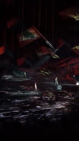 The video shows a stage performance with three individuals dressed in what appears to be traditional or ceremonial clothing. They are positioned on a multi-tiered stage that is illuminated with dramatic lighting, creating a sense of depth and shadow. The stage floor is covered in a textured surface that reflects light, resembling water or a rocky terrain. Above the stage, several large, rectangular objects are suspended, seemingly at different angles, some of which appear to be screens displaying images or projections. The overall ambiance is dark and theatrical, with a focus on the figures and their placement within the elaborate stage design. One figure in the center is raising their arm, possibly in a gesture or during a performance. The other two figures are standing further back on the stage. The lighting is a key element, with spotlights illuminating the performers and casting long shadows. The background suggests a set designed to evoke an ancient or dramatic setting.
