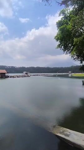 A tranquil lake scene unfolds with a floating structure, likely a small house or cabin on pontoons, in the middle distance. The structure has a reddish-brown roof and appears to be anchored to a series of connected floating platforms. The lake's surface is calm, reflecting the cloudy blue sky and a distant, tree-covered shoreline. The perspective is from a slightly elevated position near the water's edge, with a concrete structure visible in the foreground. The overall atmosphere is serene and natural, with lush green foliage visible on the right side of the frame.