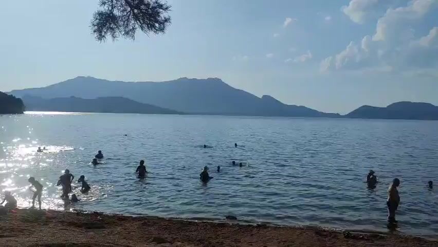 The video shows a wide shot of a lake or sea with a sandy shore in the foreground. The water is calm with gentle ripples reflecting the sunlight. Several people, mostly children and adults, are in the water, some wading, others swimming or playing. On the shore, a few people are visible, one adult standing near the water's edge, and a group of children interacting near the sand. In the background, a silhouetted mountain range stretches across the horizon under a clear blue sky with scattered white clouds. The lighting suggests it is daytime, likely late afternoon due to the angle of the sun creating shimmering reflections on the water.