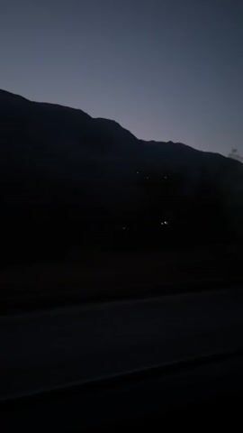 The video is a horizontal, medium shot of a darkened mountain range at twilight. The sky is a deep, muted blue, and the mountains are silhouetted against it. There are a few faint lights visible on the mountainside, possibly from houses or buildings. The foreground of the video shows a dark road, with the edge of it visible at the bottom. There is some slight motion blur, suggesting the video was filmed from a moving vehicle. The overall mood is quiet and serene.