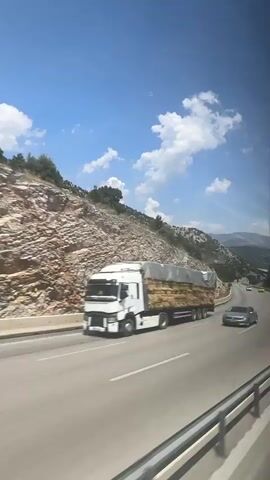 A wide shot captures a highway curving through a mountainous landscape under a bright blue sky dotted with white clouds. A truck covered with a tan tarp is traveling on the highway. The truck is loaded with large, rectangular objects, possibly bales of hay. Another vehicle, a white van, passes by. In the distance, two yellow cars are visible on the highway. The mountainside is steep and rocky, with sparse green vegetation. A highway sign with a camera icon is visible next to the road. The shot is taken from a moving vehicle, likely a bus or car, as indicated by the blurred foreground and the perspective.