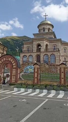 The video begins with a view of a large, ornate building with a golden dome, likely a church, situated in front of a steep, forested mountainside under a partly cloudy sky. A brick archway and a decorative fence with pillars are visible in the foreground. The camera then pans to the left, revealing a gate with a small guardhouse and a sign with an arrow pointing inward. Further along, a gray sedan is parked on the right side of the road. The camera continues to pan, showing more of the mountainside, which is densely covered in trees. Several informational signs are posted on a corrugated metal fence. The road curves to the right, and more cars, including a white sports car and a dark SUV, are visible in the distance. The video ends with a wider shot of the road and parked cars with the mountainside in the background.