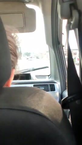 The video shows the interior of a car from the perspective of a passenger in the back seat. The view is directed forward through the windshield. The sun is bright, creating glare. In the foreground, the headrest and shoulder of a dark-colored car seat are visible. The back of a person's head and neck, with dark hair, is also in the foreground, facing away from the camera. The dashboard of the car is visible, including an air vent and part of the radio. The windshield wipers are in their resting position. Through the windshield, a street scene can be seen with several cars in the distance, appearing to be moving. The sky is bright and appears to be daytime. A seatbelt is visible on the right side of the frame.