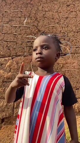 The video shows a young child standing in front of a mud brick wall. The child is wearing a dress with red, white, and blue stripes. The child is looking to the left and has their right hand raised with two fingers extended. The background wall appears to be made of packed earth and straw, with visible horizontal supports. The lighting suggests it is daytime outdoors.